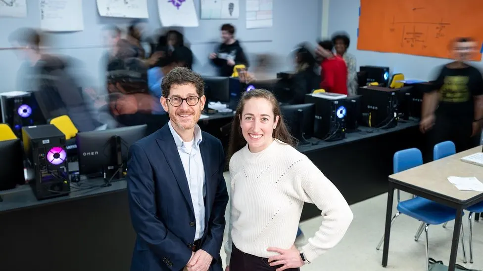 Brian Jacob and an educator smile for a photo in a computer lab classroom, with students actively working in the background.