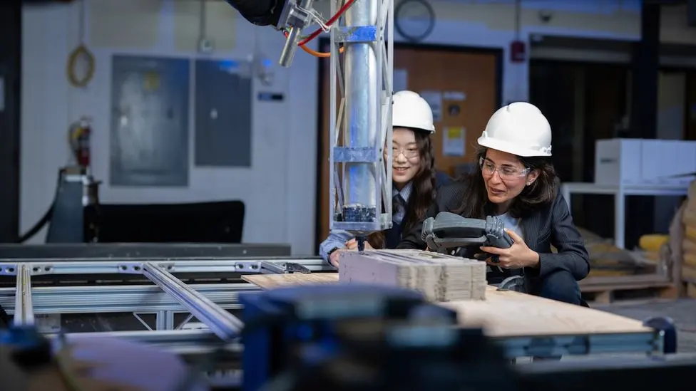 Two researchers in hard hats observing a 3D-printed concrete sample on a printer gantry in a lab