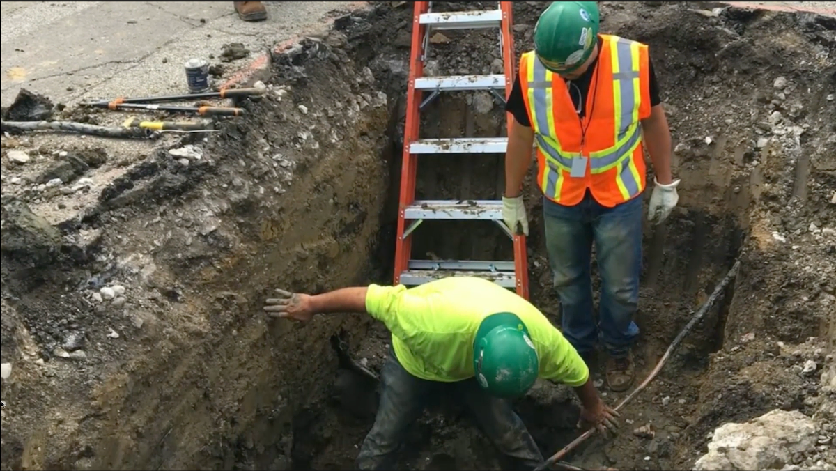 Two construction workers in hard hats stand inside a deep excavation trench, with an extension ladder leaning against the dirt wall behind them