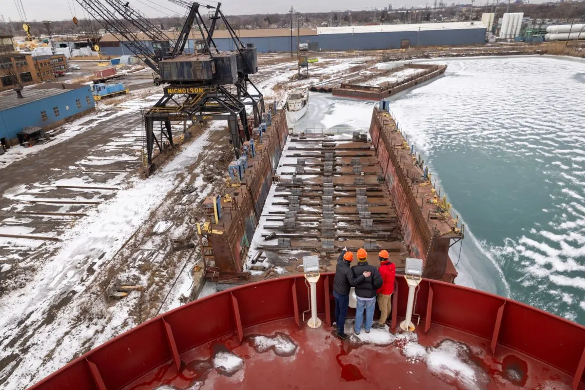 Three people stand on the deck of a shipping vessel in a harbor