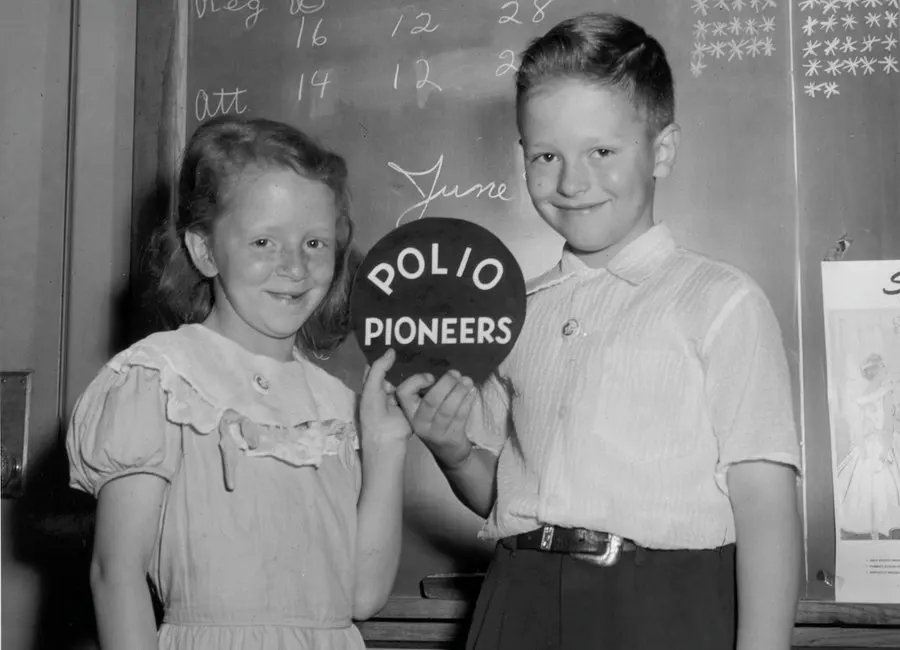 A black and white photo of two school children standing in front of a blackboard while holding a sign that says Polio Pioneers