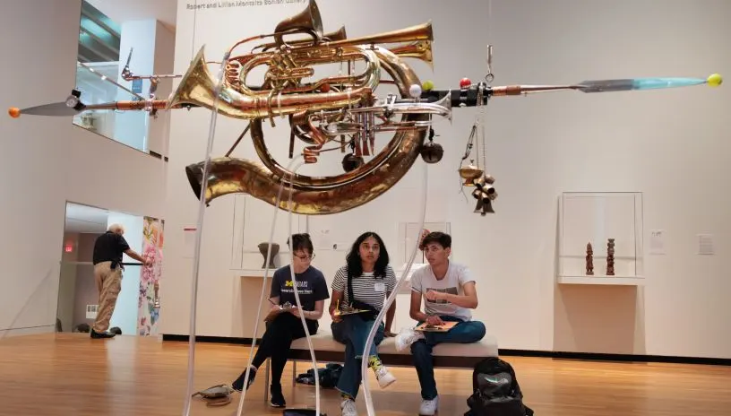 Students sitting under an art installation made of musical instruments at UMMA