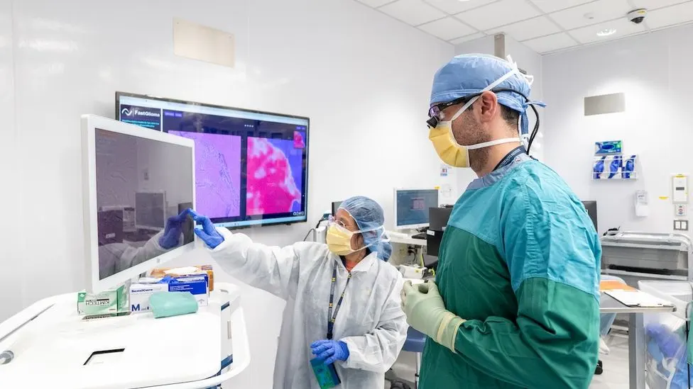 Two medical professionals in surgical gowns, masks, and gloves review brain tissue imaging displayed on a monitor showing the FastGlioma AI software in a clinical setting